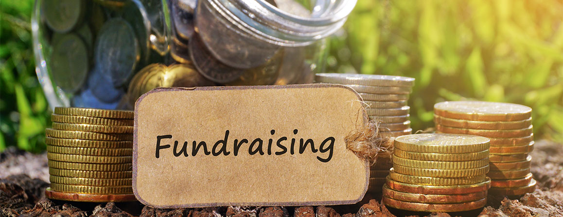  A close up of a fundraising jar with a tag on it that says 'Fundraising'. The jar is filled with pennies, nickels, dimes, and quarters. There are also stacks of pennies, nickels, dimes, and quarters next to the jar.